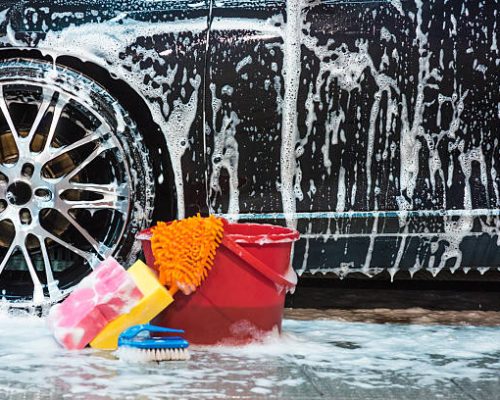 Bucket with sponges and brush with lots of foam next to car tire.